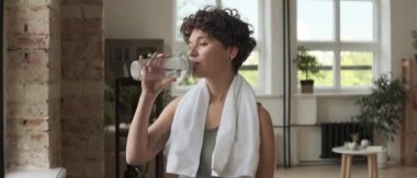 Boynunda havluyla pencerenin önünde duran ve yoga antrenmanından sonra su şişesinden su içen neşeli genç bir kadın. Anamorfik mercekle çekilen görüntü