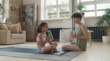 Kısa kıvırcık saçlı neşeli bir annenin ve kızının spor giyim kuşağında yoga yaptıktan sonra sohbet ederken görüntüsü.
