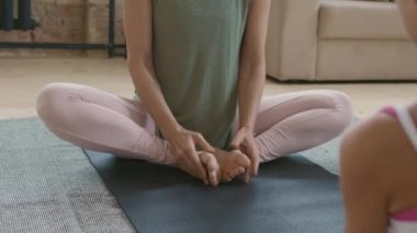 Spor kıyafetleriyle Bhadrasana 'da yoga minderinde oturan ve küçük kızına evde uzanırken talimatlar veren tanınmamış genç bir kadının orta bölüm çekimi.