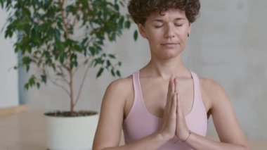 Nilüfer pozisyonunda oturmuş dua eden ve kameraya bakan sakin genç kadının portresini kaldır.
