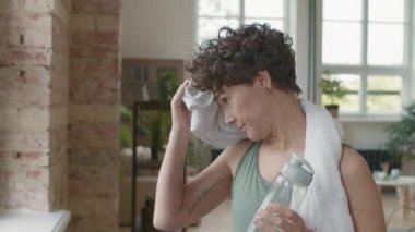 Yavaş yavaş, kısa kıvırcık saçlı, elinde su şişesi olan ve alnından ter silerken pencereden dışarı bakan yoga antrenmanından ya da egzersizden sonra dinlenen yorgun genç bir kadının portresi.