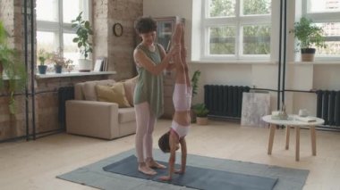 Kıvırcık saçlı, kızının yoga minderinde amuda kalkmasına yardım eden, sonra da beşlik çakan neşeli genç bir anne.