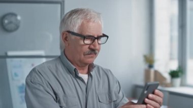 Orta ölçekli bir portre, yaşlı ofis müdürünün gözlük taktığı, akıllı telefondan mesaj attığı, sonra da kameraya baktığı görüntüleri içeriyor.