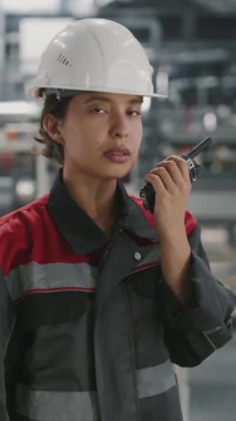 İşyerinde kameraya bakan telsizli yetenekli, melez kadın fabrika işçisinin dikey yavaş portresi.