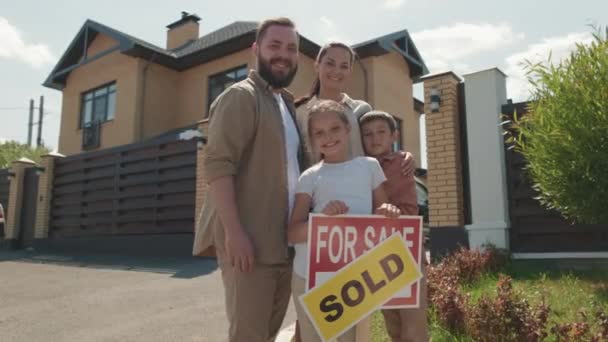 照片上 快乐的男人 女人和可爱的孩子们站在新房子前 摆出出售的姿势 并在上面贴上标牌 — 图库视频影像