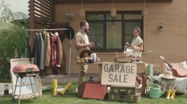  Yazın banliyö evlerinin ön bahçesinde neşeli bir adamla kadının sohbet edip garaj satışına hazırlanırken sonra da kameraya bakıp gülümserken elde çekilmiş geniş bir görüntü.