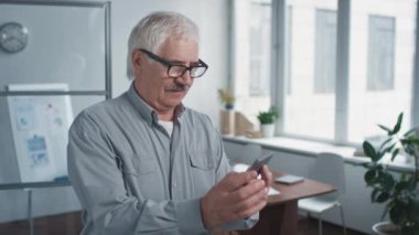 Modern üst düzey ofis çalışanı akıllı telefondan bir şeyi kontrol ediyor sonra kameraya bakıyor, belden yukarı, portre çekimi görüntülerine bakıyor.