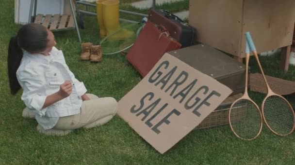 Podręczne Śledzenie Dziewczyny Siedzącej Zielonym Trawniku Piszącej Sprzedaż Garażową Kartonie — Wideo stockowe