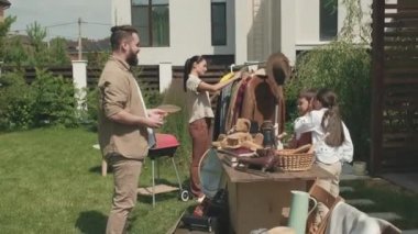  Erkek ve kızın ön bahçede garaj satışı yaparken görüntüsü. Masanın arkasında duruyorlar ve tenis raketi tutan sakallı adamla konuşuyorlar. Elbise askısında asılı ceketlere bakan genç bir kadın.