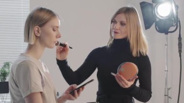 Güzel kadın MUA 'nın yavaşça fırça kullanırken ve cep telefonlu güzel kadın modelin yüzüne pudra sürerken görüntüsü.