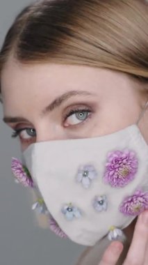 Çiçeklerle süslenmiş, kameraya bakıp poz veren yüz maskeli kadın modelinin dikey aşırı yakın çekimi.