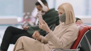 Ortadoğulu genç bir adam arka planda müzik dinlerken, orta boy, koruyucu maskeli yetişkin beyaz kadın kalkış salonunda oturmuş akıllı telefondan video görüşmesi yapıyor.