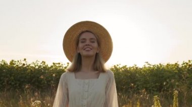 Ay çiçeği tarlasında kameraya doğru yürüyen, yaz elbisesi ve hasır şapkalı güzel beyaz bir kadının yavaş çekim portresi.