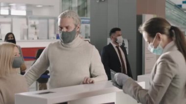 Yetişkin, beyaz bir çiftin kayıt gişesinde durduğu orta boy bir fotoğraf. Kadın servis elemanı, biniş kartı ve koruyucu maskeler takan herkesin belgelerini kontrol ederken.
