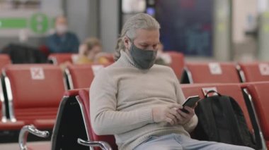Coronavirus sırasında kalkış salonunda uçuş beklerken akıllı telefon kullanan koruyucu maskeli yaşlı beyaz adamın orta boy yakın çekimi.