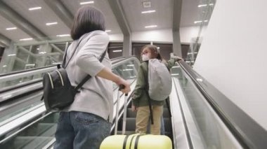 Düşük açılı yavaş çekim. İki küçük kız ve büyük bavul taşıyan bir kadın havaalanında yürüyen merdivende yürüyor.