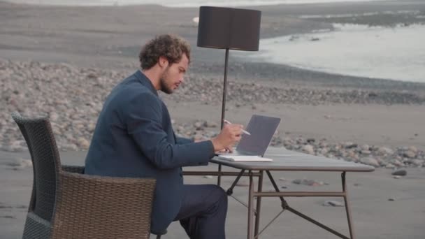 穿着蓝色夹克的商人坐在蓝色大海前的办公桌前 在笔记本电脑上工作 — 图库视频影像