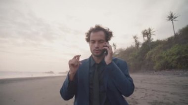 Tropikal bölgede boş bir plajda çalışan yakışıklı iş adamının telefon görüşmelerini gösteren drone görüntüleri.