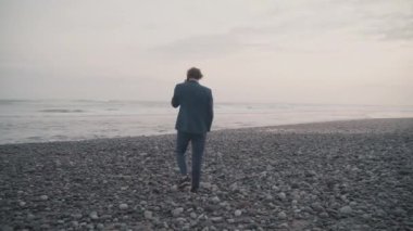 Bali 'ye seyahatte başarılı bir iş adamının insansız hava aracı görüntüsü, izole okyanus kıyısında yürürken telefon konuşması.