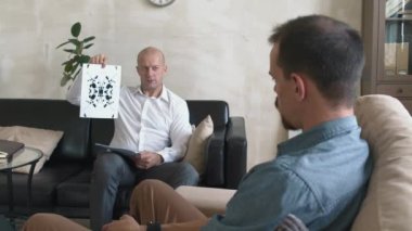 Orta yaşlı erkek psikoterapistin seans sırasında sandalyede oturan genç sakallı adama Rorschach testi yaparken görüntüsü.