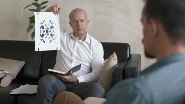 セッション中に男性クライアントにRorschachテスト写真を示す中年男性精神分析家の追跡 — ストック動画