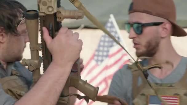ライフルで狙いと射撃を練習している軍の衣装を着た若い白人男性の中近接映像を追跡し スコープを見ながら話し 米国の国旗が背景に平らになる — ストック動画