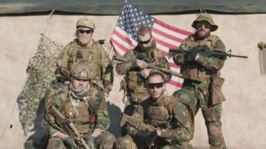 Kamuflajlı genç Amerikan istihbarat görevlilerinin orta boy portresi. Arka planda Amerikan bayrağı ile birlikte duran tüfekler.