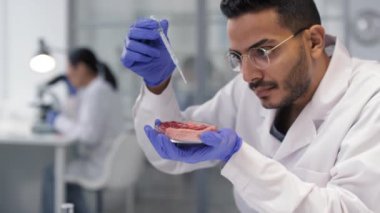 Beyaz laboratuvar önlüklü ciddi bir ortadoğu bilim adamının, petri kabından alınan yapay et örneğini inceleyip, üzerinde damlatan su dağıtıp tepkimeyi beklemesi.