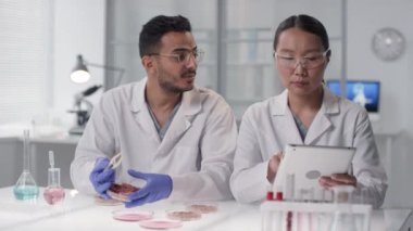 Gülümseyen mikrobiyoloji bilimadamlarının, laboratuvar yetiştirilmiş et örneklerini dijital tabletle inceleyen ve kameraya bakan belden aşağısı ağır ağır portresi.