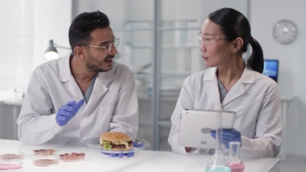 多民族科学者の腰を上げながら 白現代研究室のテーブルに腰を下ろして 研究室で育てた肉でバーガーを検査し議論する — ストック動画