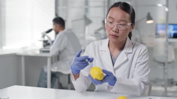 現代の研究室で顕微鏡下の物体を観察しながら 人工ビタミンを注射器でレモンを注入する女性アジアの科学者のスローモーション腰のアップ — ストック動画