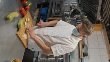 Koronavirüs sırasında restoranın mutfağında durup, kabak keserken sağlıklı yemek pişiren Kafkasyalı şefin yavaş çekimde dikey görüntüsü.
