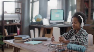 Orta ölçekli, neşeli, enerjik afro çocuğun dijital tablet kullanırken ve oturma odasındaki masada kulaklıkla müzik dinlerken ablası arka planda koltukta otururken.