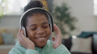 Yavaş çekimde neşeli afro kızın kulaklıkla müzik dinlemesi evde şarkı söylemesi ve şarkının tadını çıkarması.