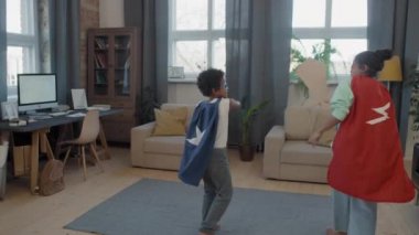 Tracking full shot of two mixed-race siblings in handmade superhero costumes fighting for fun with carton swords in living room, having fun together