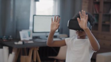 Parlak oturma odasında duran, görünmez şeylere dokunan, kulaklıklı melez bir çocuğun orta boy yakın çekim görüntülerini izliyorum.