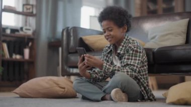 Oturma odasındaki halının üzerinde oturmuş akıllı telefondan video görüşmesi yapan, sohbet eden ve gülen neşeli afro çocukla dolu bir fotoğraf.