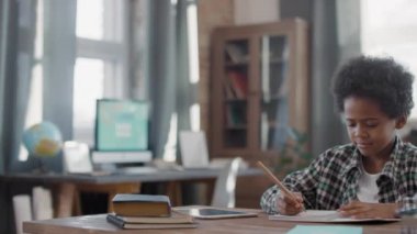 Oturma odasında oturan, ders çalışan, not defterine yazan sevimli afro çocuğun belini bükme görüntüleri.