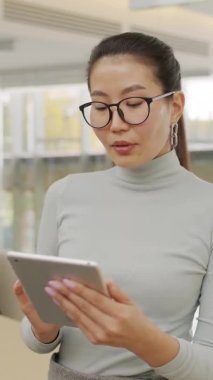 Dijital tablet tutan ve kameraya bakan Asyalı güzel iş kadınının dikey yavaş çekimde orta boy portresi.
