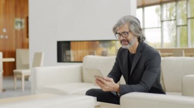 Orta yavaş çekimde, siyah ceketli gülümseyen yaşlı bir adamın portresi dinlenme yerindeki deri koltukta dinleniyor, kahveyi yudumluyor ve dijital tablet kullanıyor.