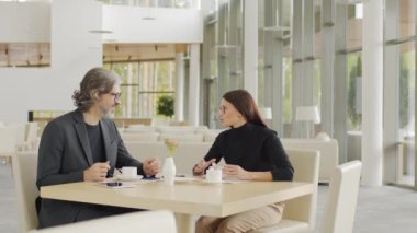 Kafkas iş ortaklarının kahve fincanlarıyla modern restoranda masada oturup iş konularını tartışırken birbirlerine gülümsedikleri orta ölçekli yavaş çekim görüntüleri.