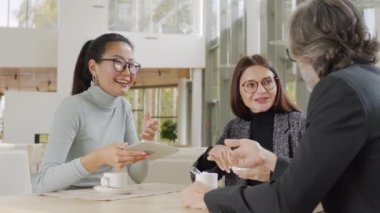 Çok ırklı iş ortaklarının yeni projeyi tartıştığı, dijital tablet kullanarak modern restoran masasında yan yana oturduğu orta ölçekli bir yakın çekim.