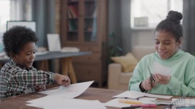 Orta boy afro çocukların sohbet ederken, kağıt işçiliğinden resimler keserken modern oturma odasında otururken.