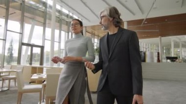 Çağdaş ofis binasının panoramik pencereli lüks lobisinde yürürken çoklu etnik çeşitlilikte gülümseyen iş ortaklarının iş tartışmalarını orta açıdan izleme