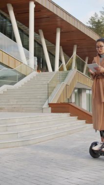 Modern ofis binasının yanında panoramik pencereli jiroskop sürerken, deve kürkü giymiş ciddi Asyalı kadının dijital tablet kullandığı dikey bir fotoğraf.