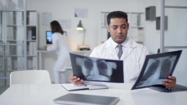 Medium portrait shit footage of professional medical worker sitting at desk in doctors office working with X-ray pictures then his colleague asking him something
