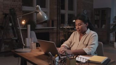Ağır çekimde, çalışkan Afrikalı Amerikalı yetişkin bir kadının portresi. Geceleri ofiste oturup dizüstü bilgisayarla çalışıyor. Sonra da gülümseyen kameraya bakıyor.