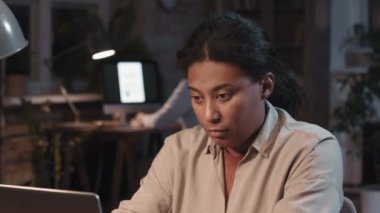 Afrika kökenli Amerikalı yetişkin bir kadının arka planda bilgisayarla yazı yazıp karanlık odada yan yana oturduğu orta-yavaş çekimde yakın plan çekimleri.