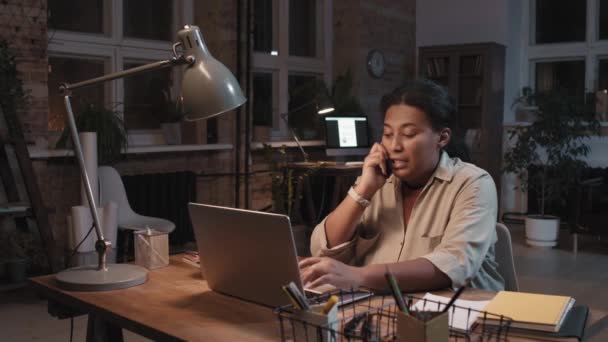 仕事中のアフリカ系アメリカ人女性が夜な夜な一人で暗いロフトオフィスのノートパソコンで仕事をしながら電話で話しています — ストック動画