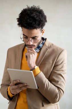 Genç ve düşünceli bir iş adamı akıllı günlük giysiler içinde kameranın önünde dikiliyor ve dijital tablet görüntülerine bakıyor.